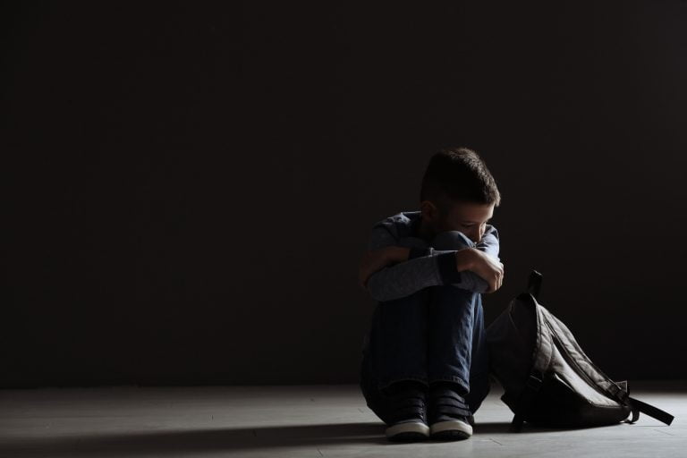 Upset boy with backpack sitting in dark room.