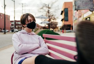 Girl sits on a bench in the city while wearing a face mask- coronavirus pandemic