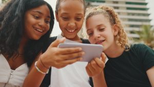 Three girls friends pre-teenage are sitting on the waterfront using mobile phone, watching videos, photos. Three teenagers on the outdoors in urban cityscape background