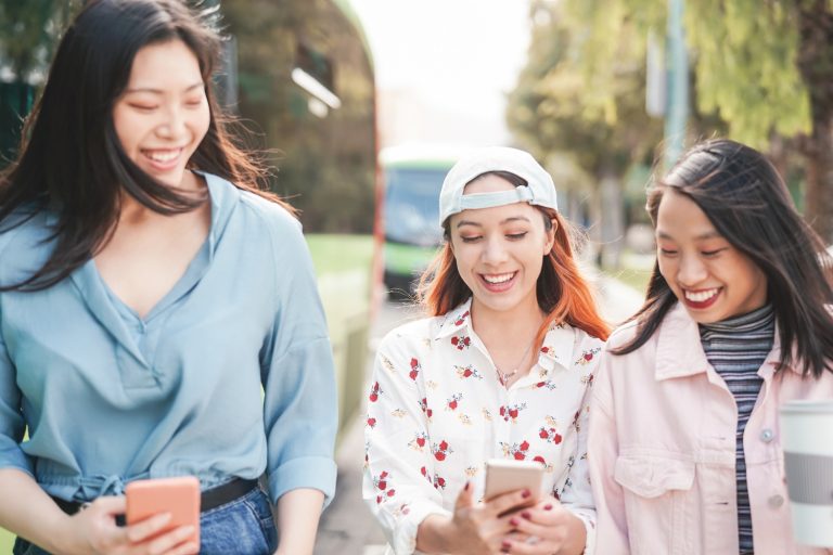 Happy Asian girlfriends walking in the city while watching on mobile smartphones - Young teen girls having fun with new social media apps - Youth millennial generation and technology concept