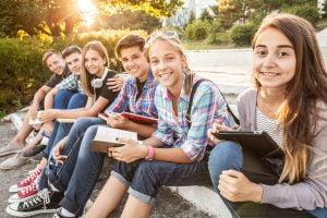group of young students with books and gadgets sit on the steps in the park