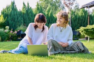 Teenage female student friends laughing sitting on grass with laptop