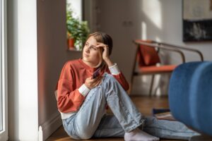 Pensive girl teenager sits on floor looks at window with smartphone in hand at home