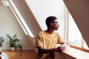 Young Man Relaxing In Loft Apartment Looking Out Of Window With Hot Drink