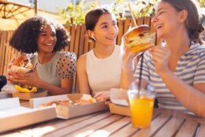 smiling teenage girls are sitting in open air cafe and eating fast food