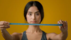 Sad upset worried Indian woman girl in yellow studio with measuring tape dissatisfied with body