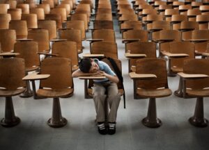 A boy sitting at the front in a hall of empty desks, with his head on his arm.