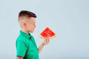 little boy with slice of watermelon in hand. isolated on blue background, copy space, in studio, profile view