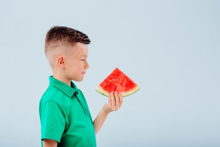 little boy with slice of watermelon in hand. isolated on blue background, copy space, in studio, profile view