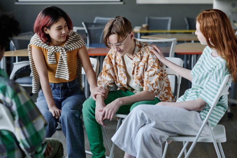 Ethnically diverse teen girls supporting sad boy during group therapy session